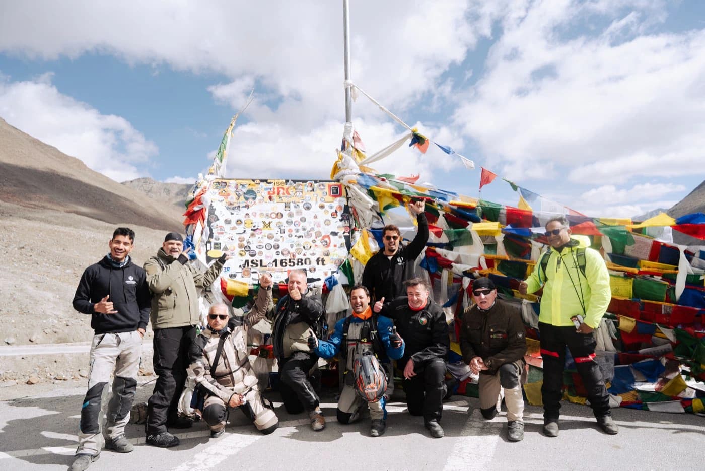 Riders and crew celebrating at the Shinku La summit board at 16,580 ft with prayer flags
