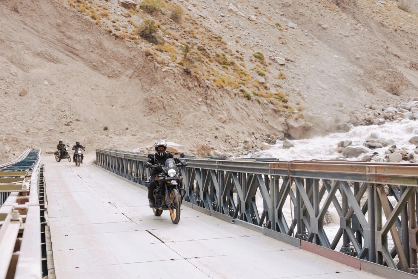 Rider on a Royal Enfield Himalayan 450 crossing a steel bridge over a glacier river in Zanskar