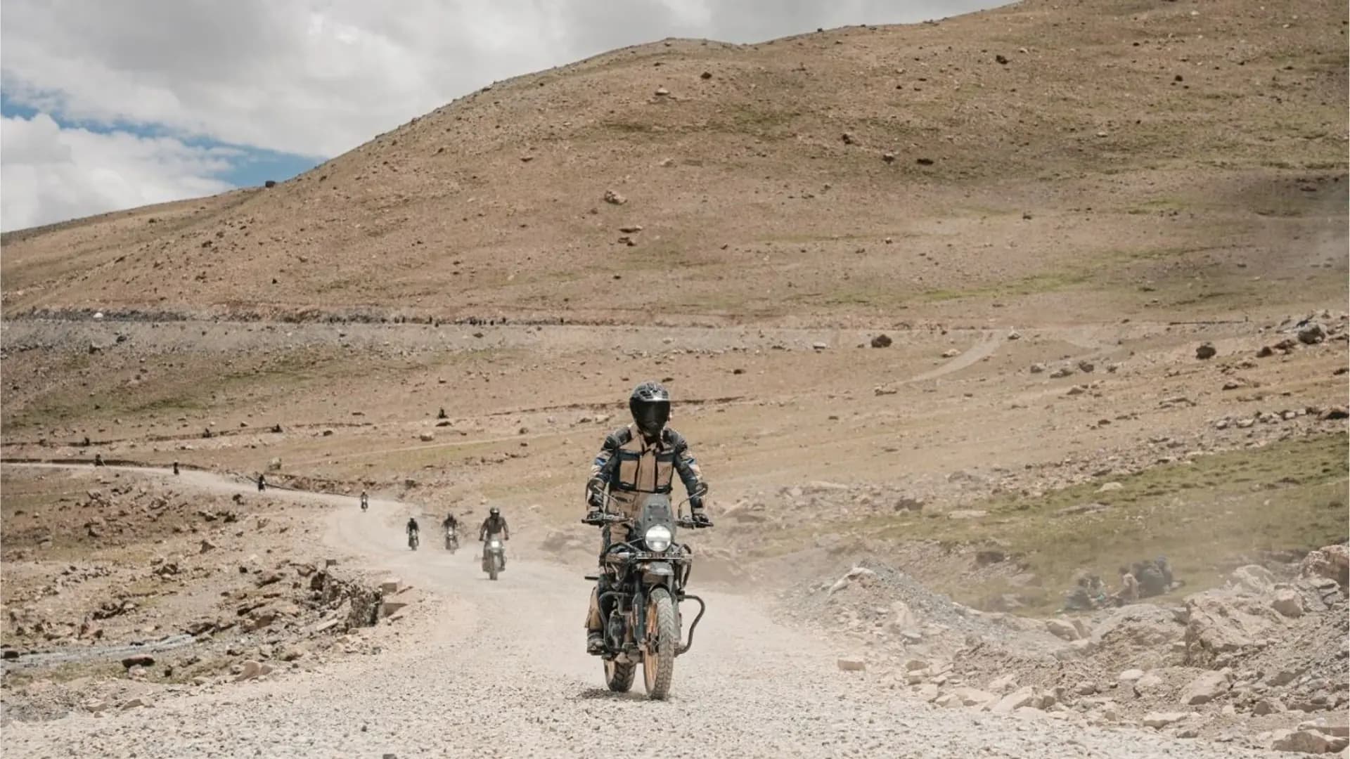 Pilote en équipement de protection sur une Royal Enfield Himalayan 450 sur une piste poussiéreuse de haute altitude au Ladakh, suivi par d'autres pilotes