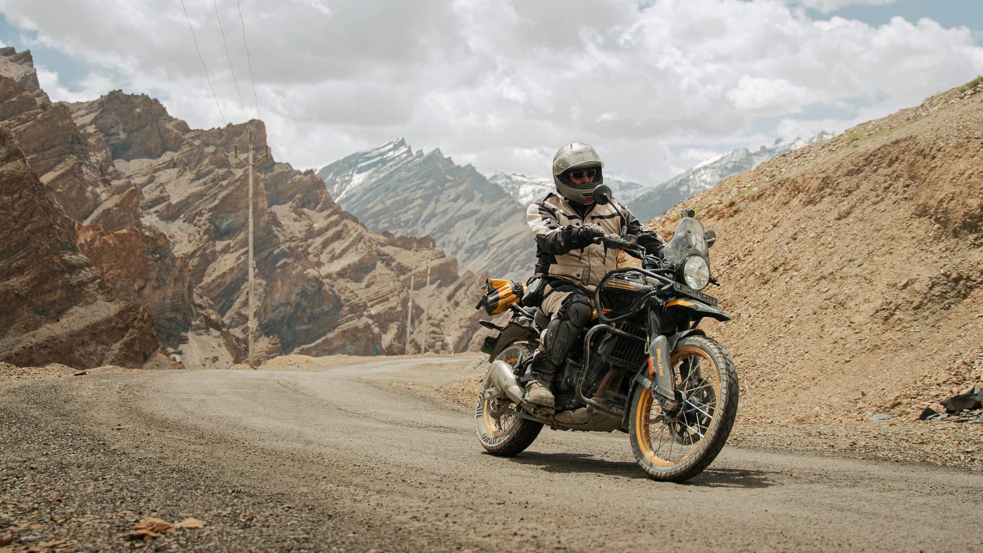 Motorcyclist riding a gravel mountain road below rocky ridges and a snow-capped peak