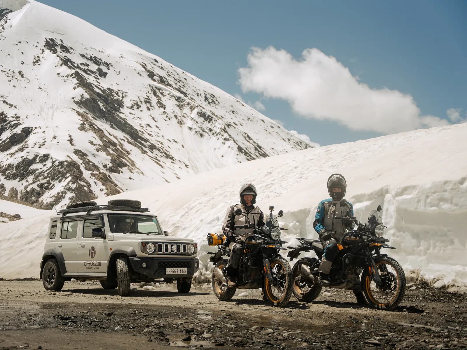 Two riders on motorcycles and a support vehicle parked beside deep roadside snow under a blue sky