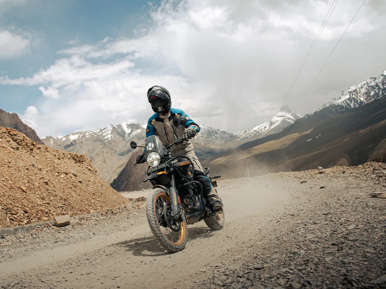 Motorcyclist riding a dusty dirt road with snow-streaked mountains behind