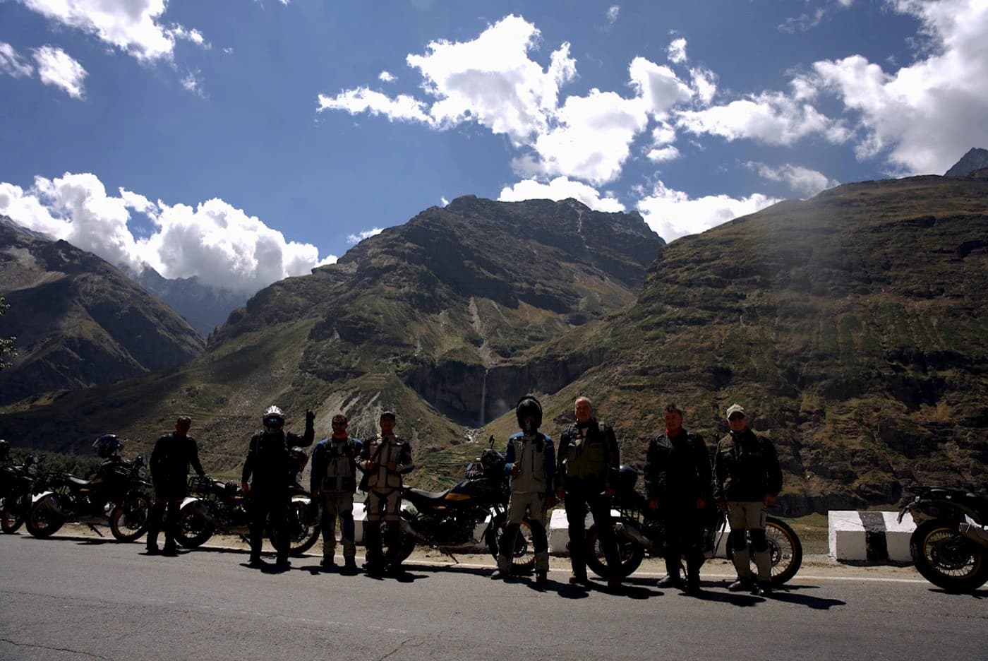 Group of motorcycle riders lined up on the road in the Lahaul valley with mountains and waterfalls behind