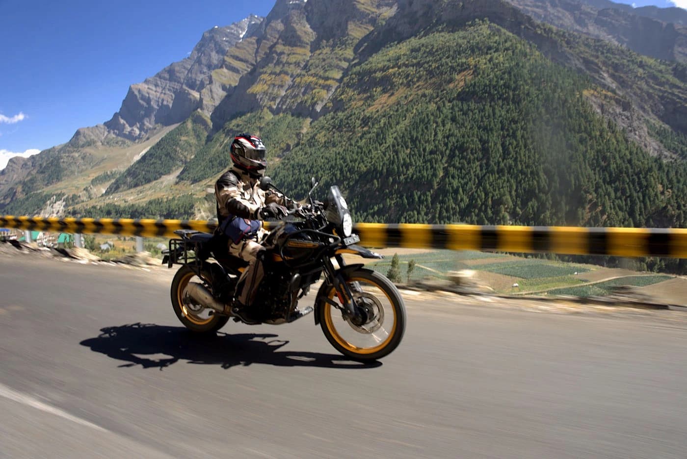 Motorcyclist riding past a crash barrier with forested Lahaul mountains behind