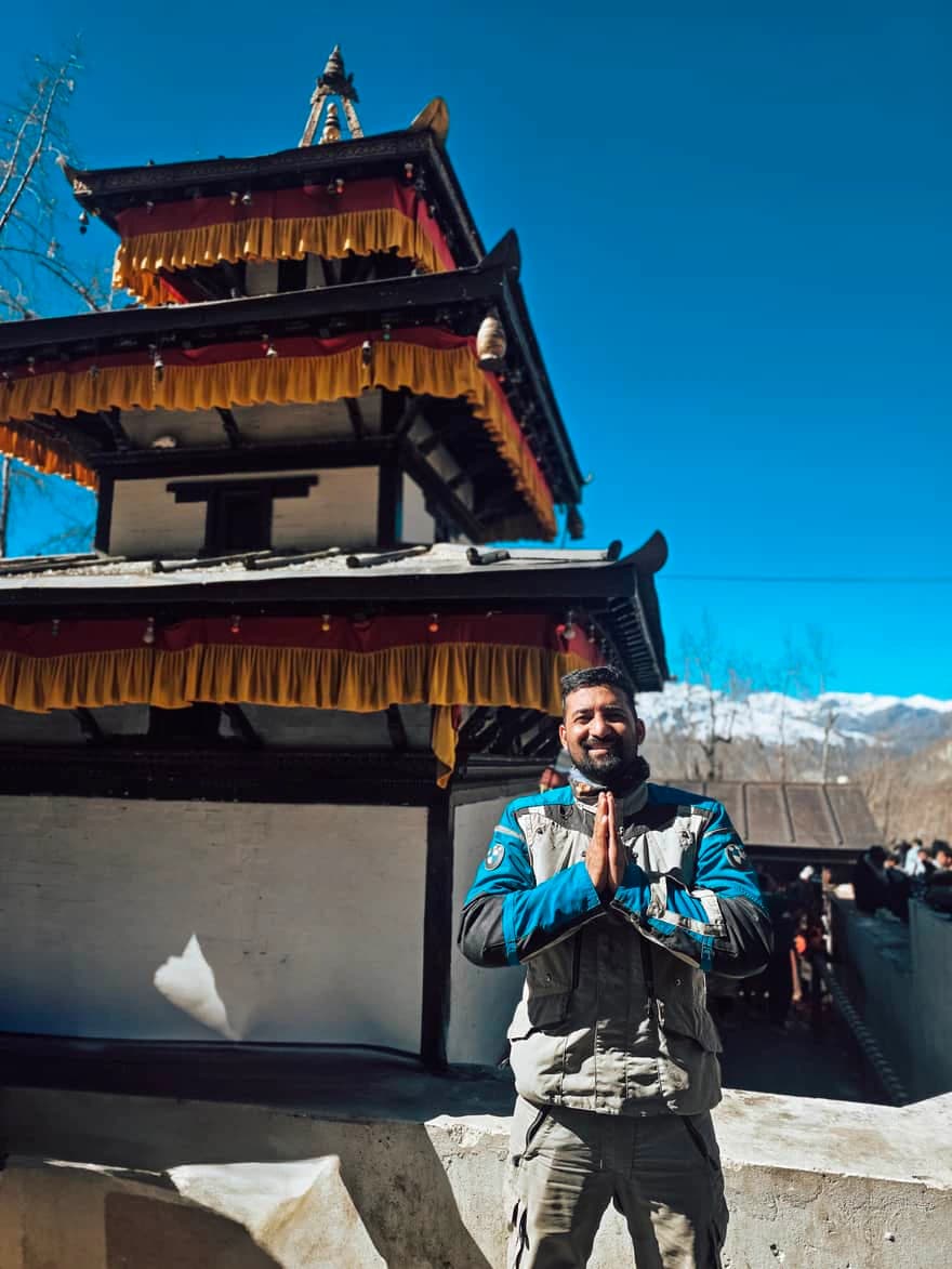Traveler standing with folded hands in front of Muktinath Temple, a sacred pilgrimage site in Nepal, with Himalayan mountains in the background.