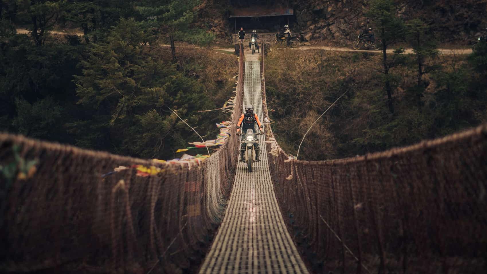 Motorbike rider crossing a suspension bridge surrounded by forested hills in Nepal.