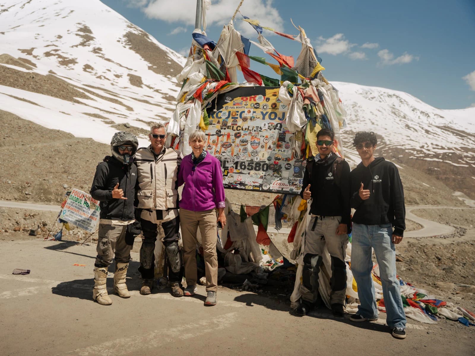 A motorcycle tour group posing at Shinkula Pass in Ladakh with colorful prayer flags and snow-covered Himalayan mountains in the background. Shinkula Pass is an important high-altitude mountain pass on the route connecting the Lahaul region with Zanskar.
