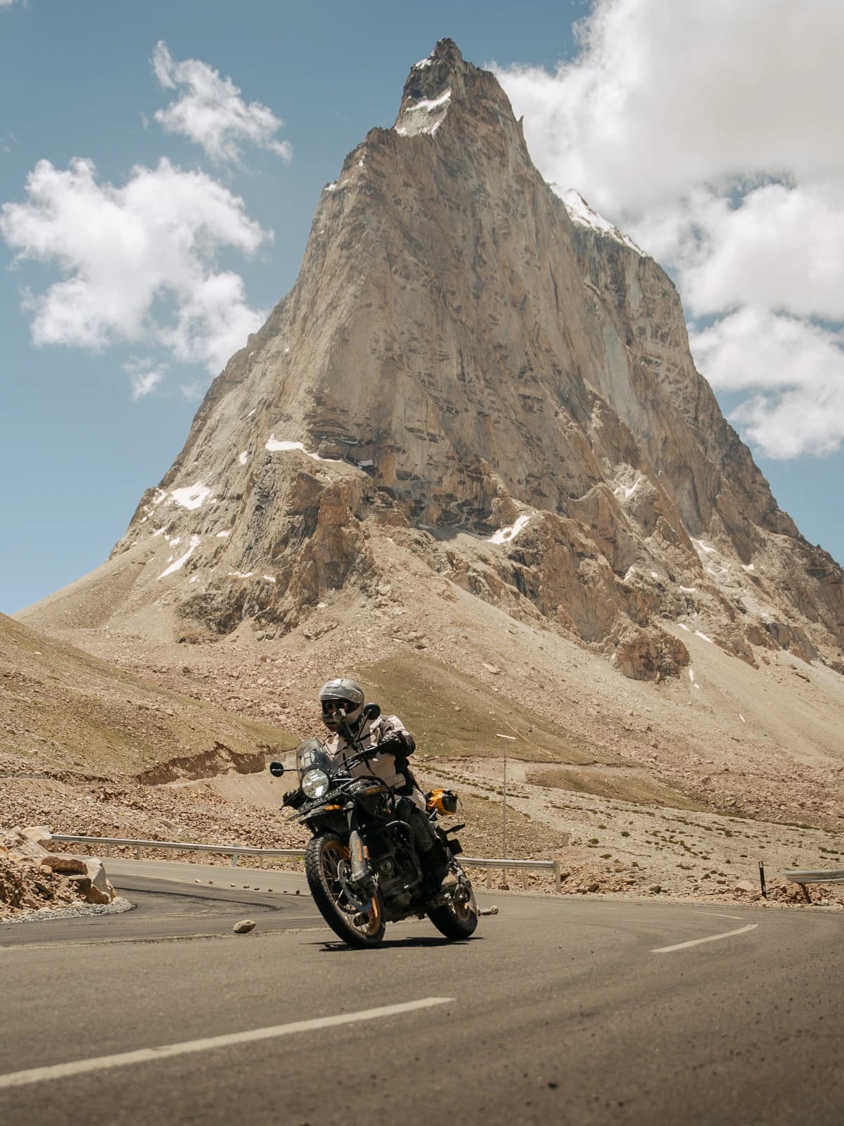A motorcycle rider on a Royal Enfield Himalayan riding through the high-altitude landscape of Gombok Rongjon in Ladakh, with a dramatic rocky Himalayan peak towering in the background.