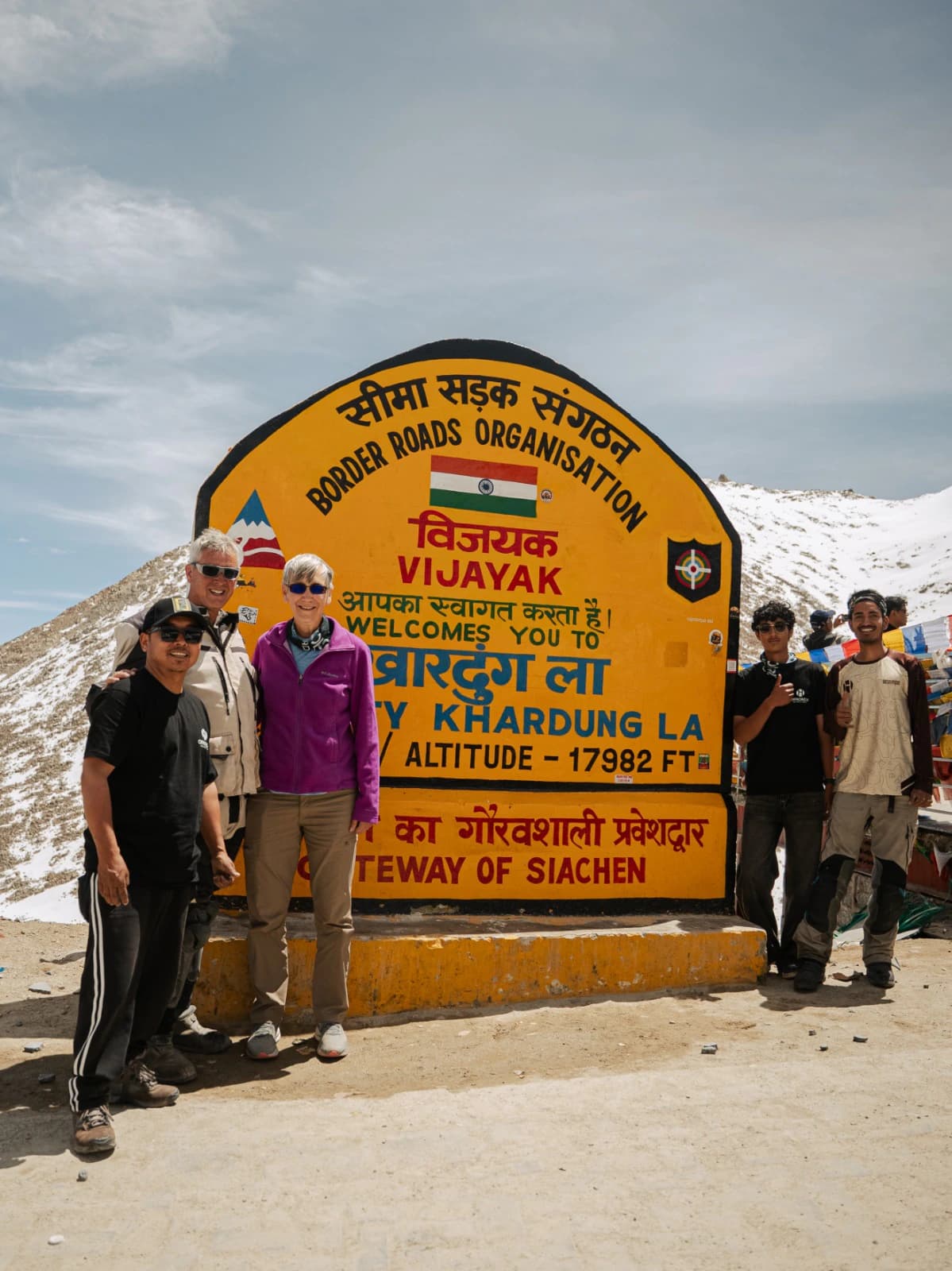 A motorcycle tour group posing beside the iconic Khardung La Pass sign in Ladakh, surrounded by snow-covered Himalayan mountains. The stop is part of an adventurous Leh Ladakh motorcycle tour through the high-altitude landscapes of the Himalayas.