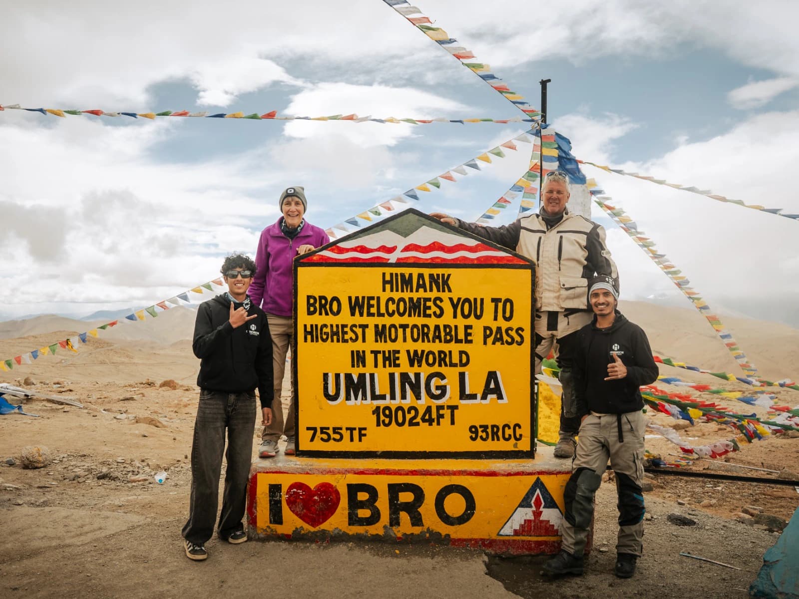 A motorcycle tour group posing at the Umling La Pass sign in Ladakh, surrounded by colorful prayer flags and the vast high-altitude Himalayan landscape. Umling La is a spectacular destination for riders exploring the remote landscapes of Ladakh.