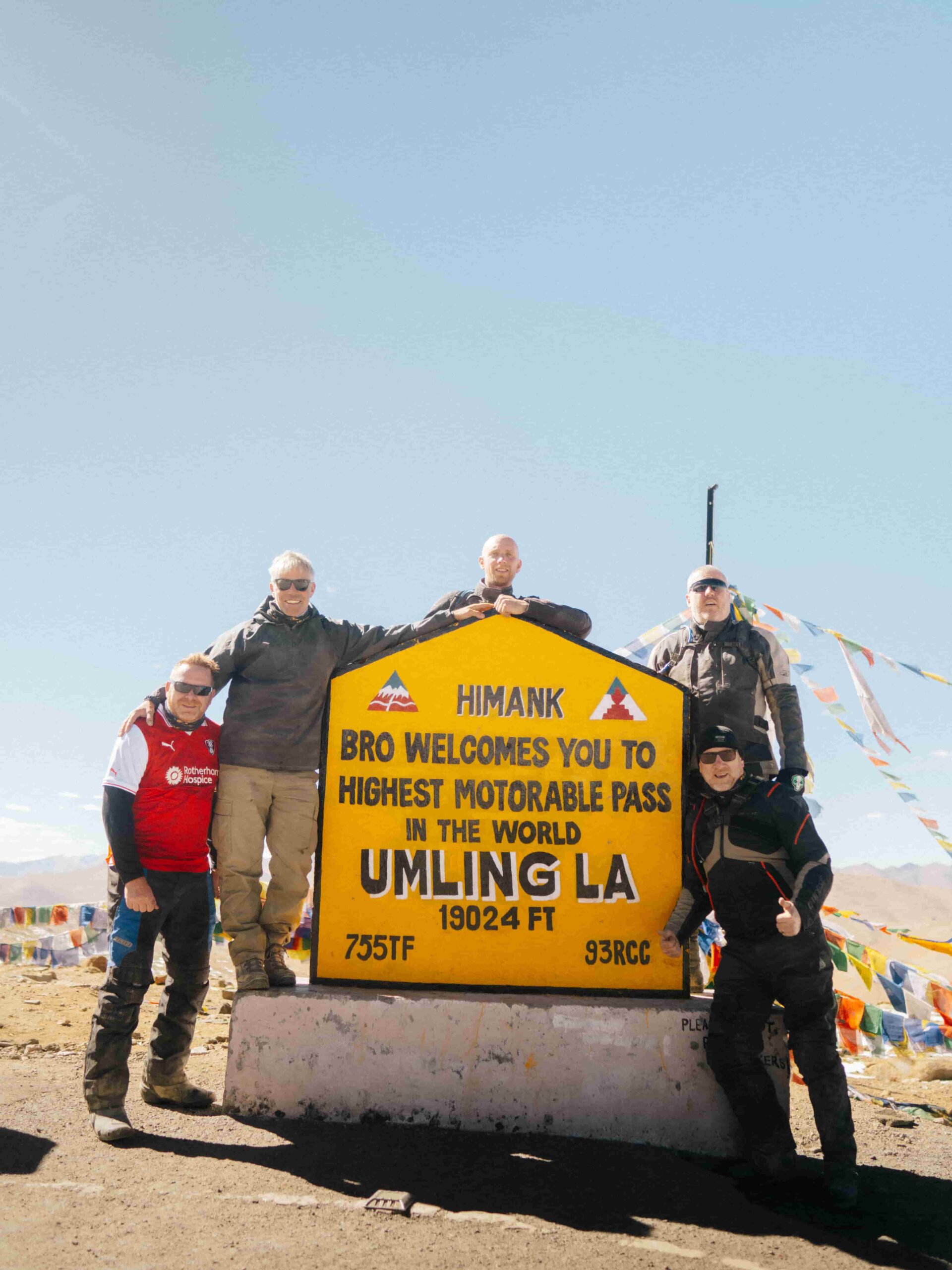 Group of travelers posing at Umling La Pass signboard, the world’s highest motorable road at 19,024 feet in Ladakh, India, under a clear blue sky.