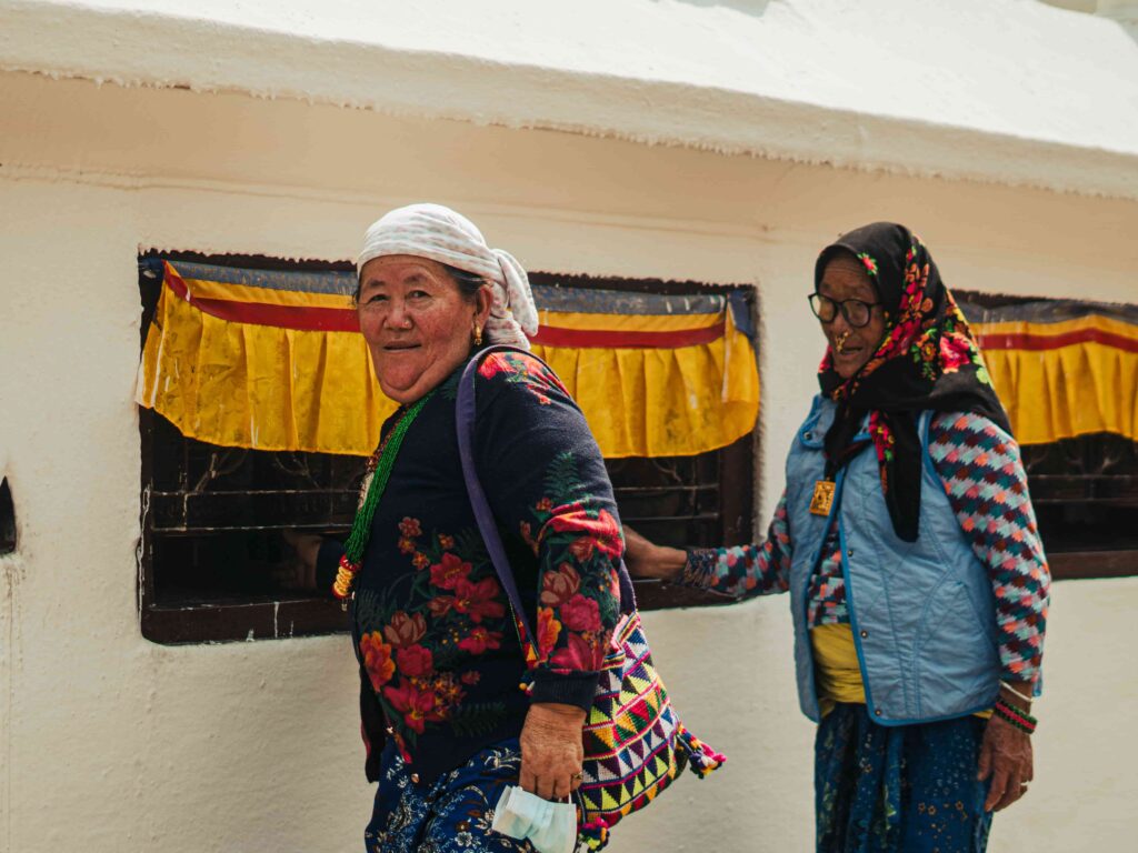 Two elderly Nepali women wearing traditional clothing walking beside a monastery wall with colorful decorations