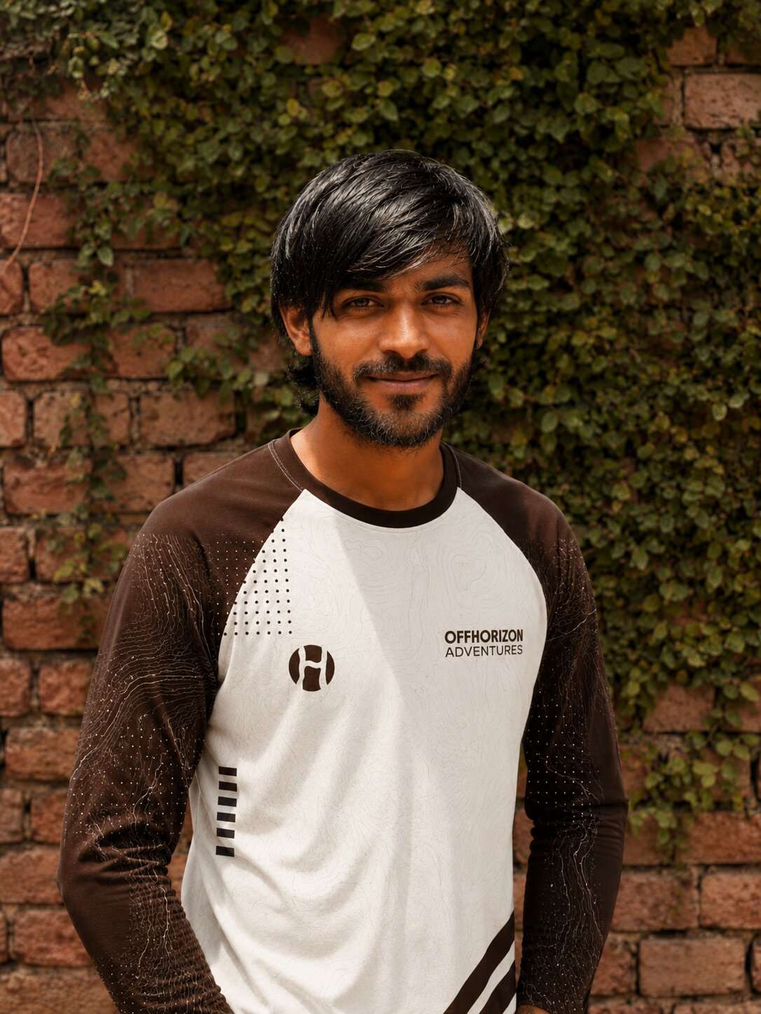 Portrait of Ajay from Offhorizon Adventures wearing branded outdoor clothing in front of a brick wall covered with green vines.