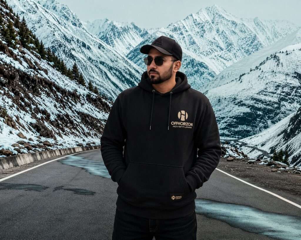 Tushar Ranta from OffHorizon Adventures standing on a mountain road surrounded by snow-covered peaks wearing a black hoodie and sunglasses.