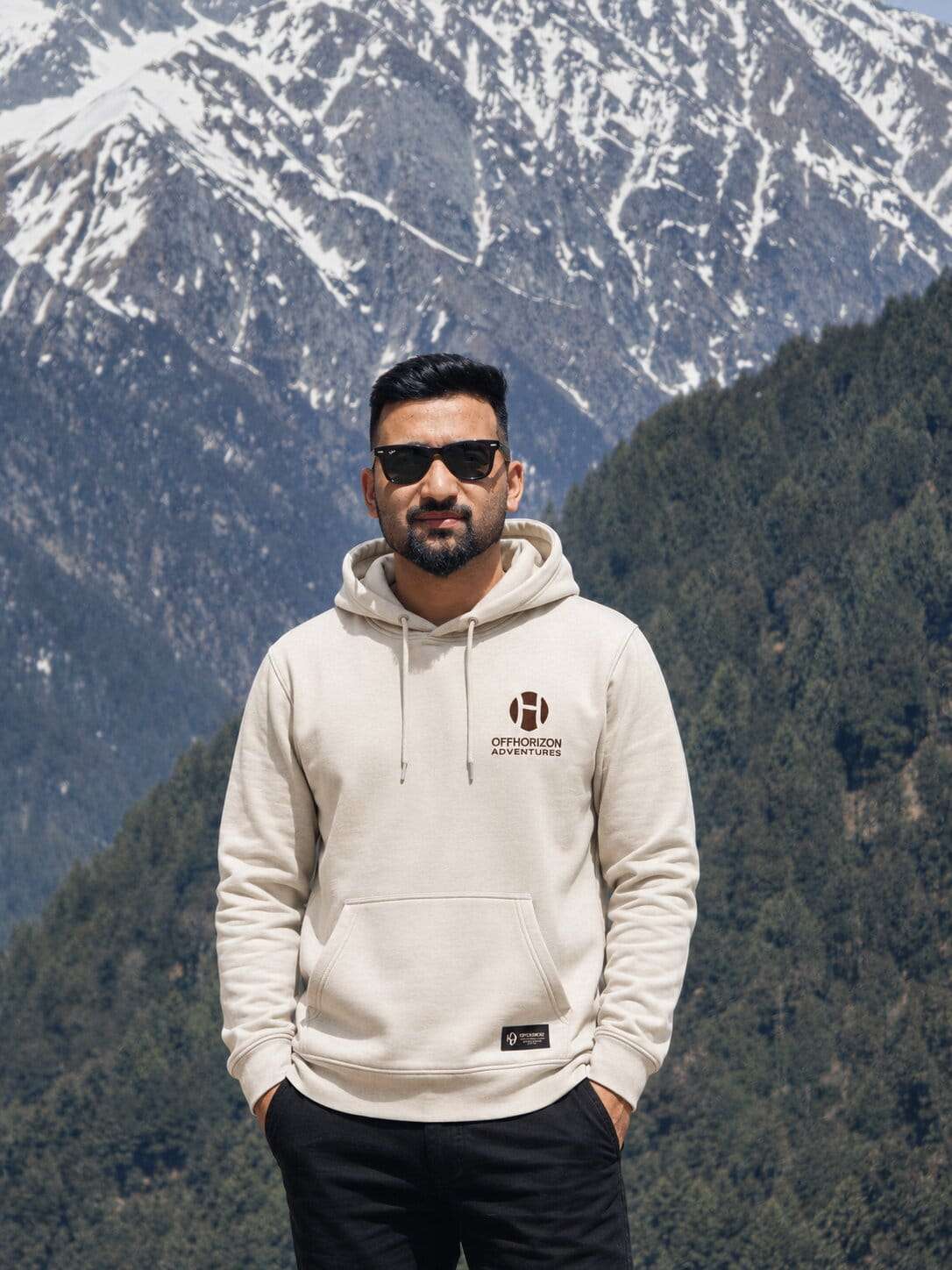 Surya Thakur of Offhorizon Adventures standing outdoors with snow-covered mountains in the background wearing a beige hoodie and sunglasses.